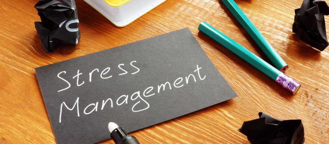 Stress Management written on grey card surrounded by office supplies on wooden desk representing workplace stress management techniques