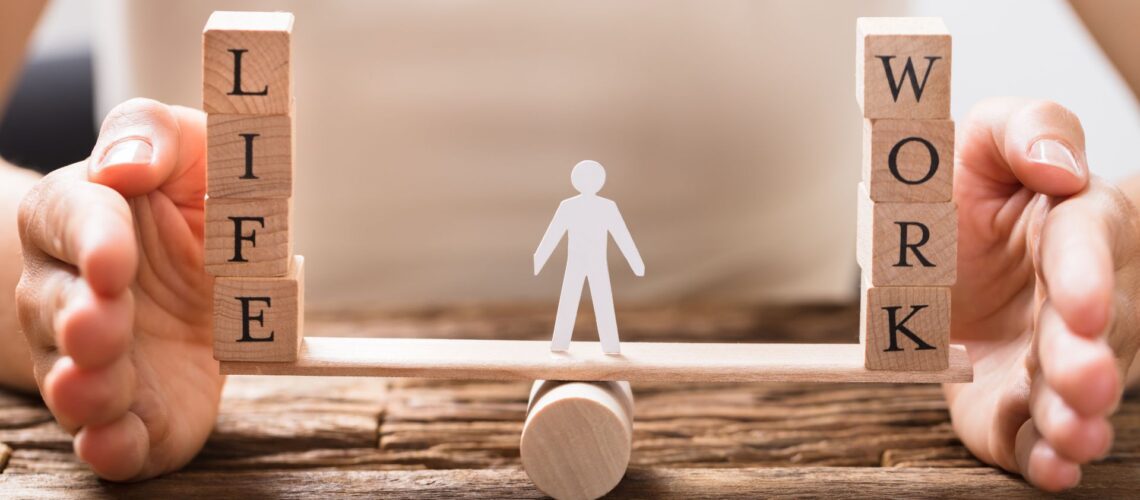 Hands protecting wooden blocks spelling LIFE and WORK balanced on a scale, representing work-life balance strategies for building resilience at work