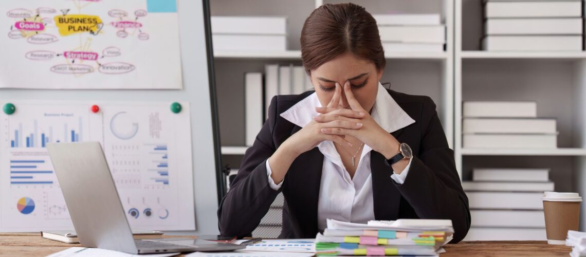 Professional woman with hands on face showing workplace stress, surrounded by business charts and laptop, illustrating barriers to building resilience at work
